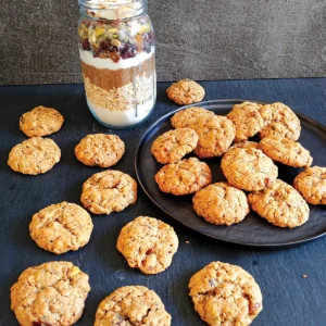 A batch of cookies arranged on a table and stacked on a dish, with a mason jar full of the ingredients at the back of the table.