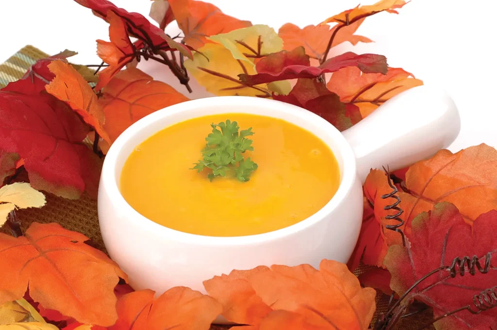 A white bowl of roasted butternut squash soup set upon a pile of autumn leaves.