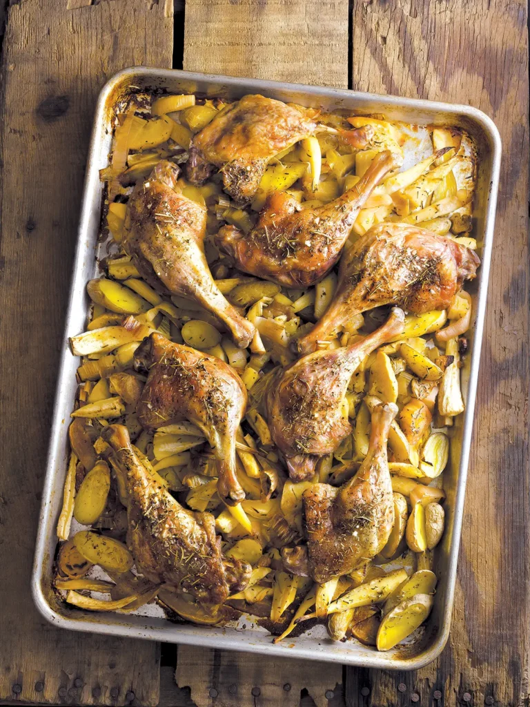 A sheet pan containing roasted duck legs with apples, parsnips and leeks set on a wooden table.