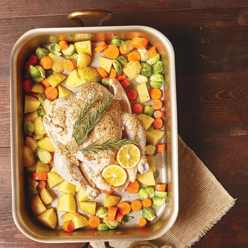 Roasting pan containing vegetables and a whole mustard coated chicken.