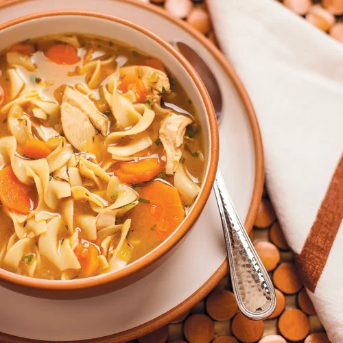 A bowl of chicken noodle soup set on a wooden placemat.