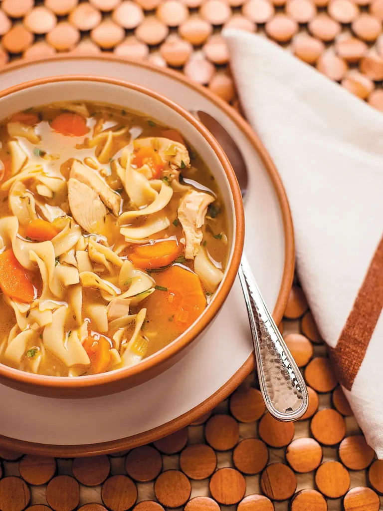 A bowl of chicken noodle soup set on a wooden placemat.