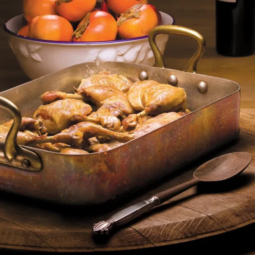 Duck confit in a roasting pan next to a bowl of persimmons.