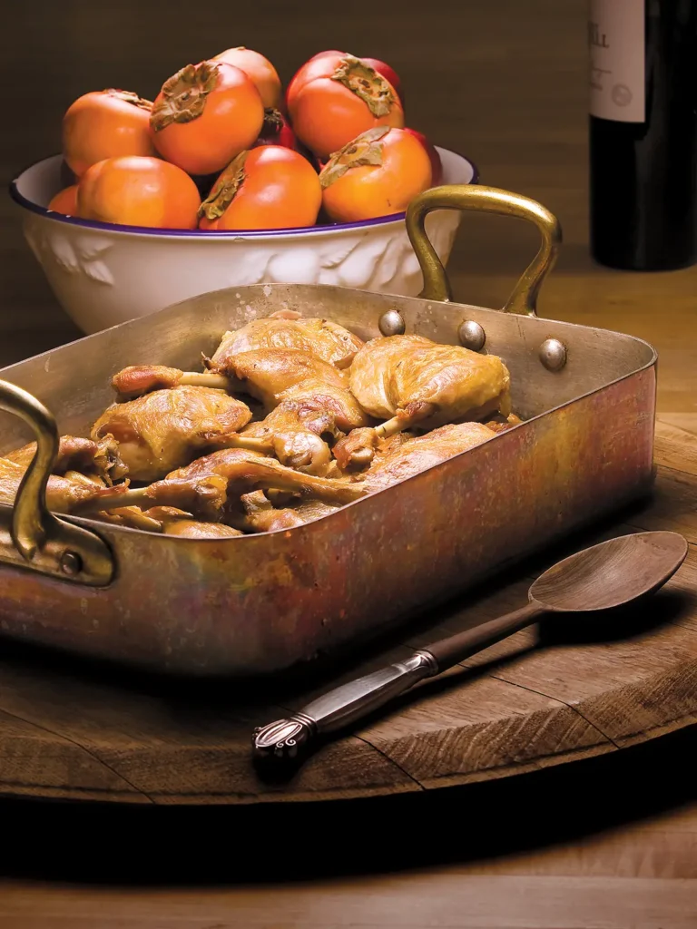 Duck confit in a roasting pan next to a bowl of persimmons.