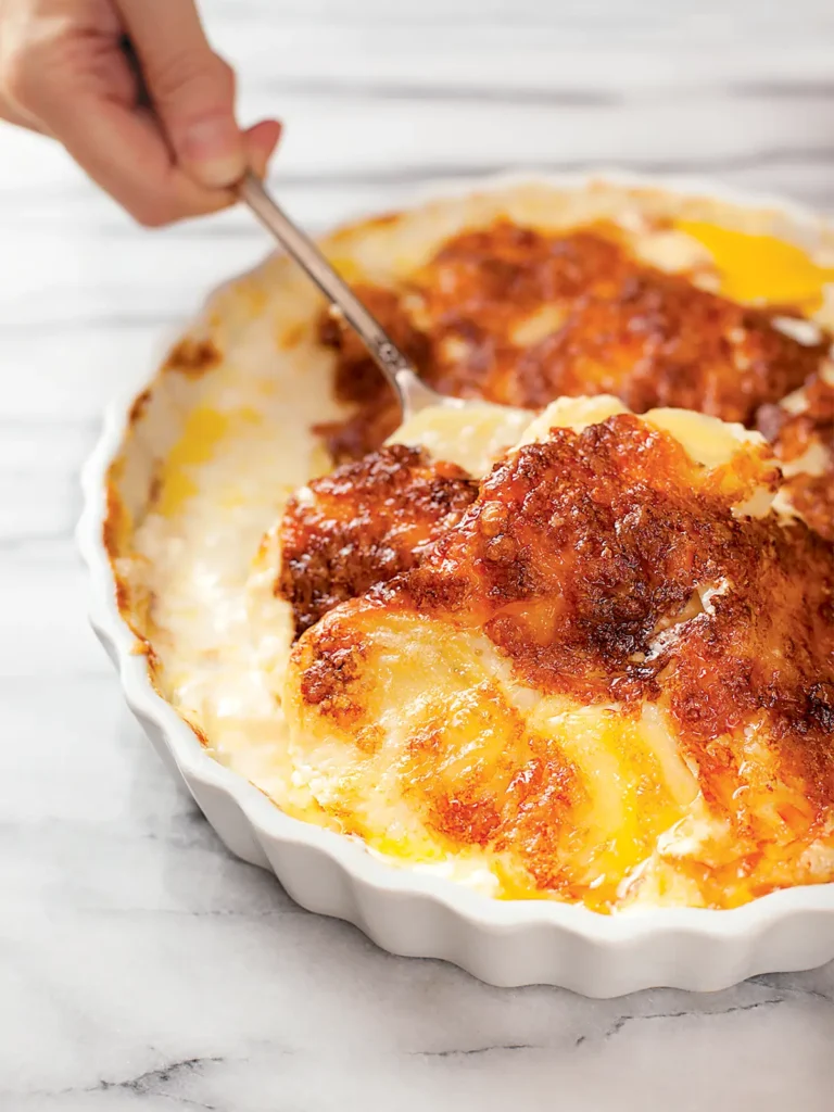 A hand with a spoon, scooping Gratin Dauphinois from a white scalloped baking dish.