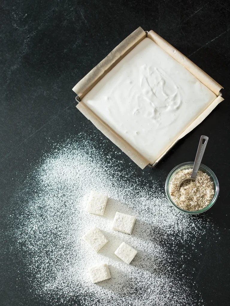 6x8 pan of homemade marshmallow next to a jar of marshmallow root and several marshmallows with powdered sugar dusting on a black granite counter top.