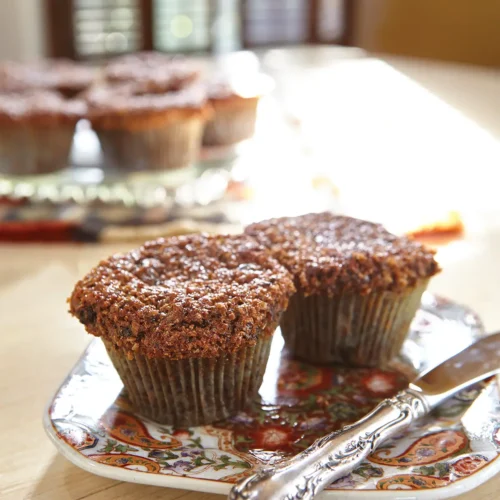 Two raisin bran muffins set on a patterned plate with a knife resting next to them.