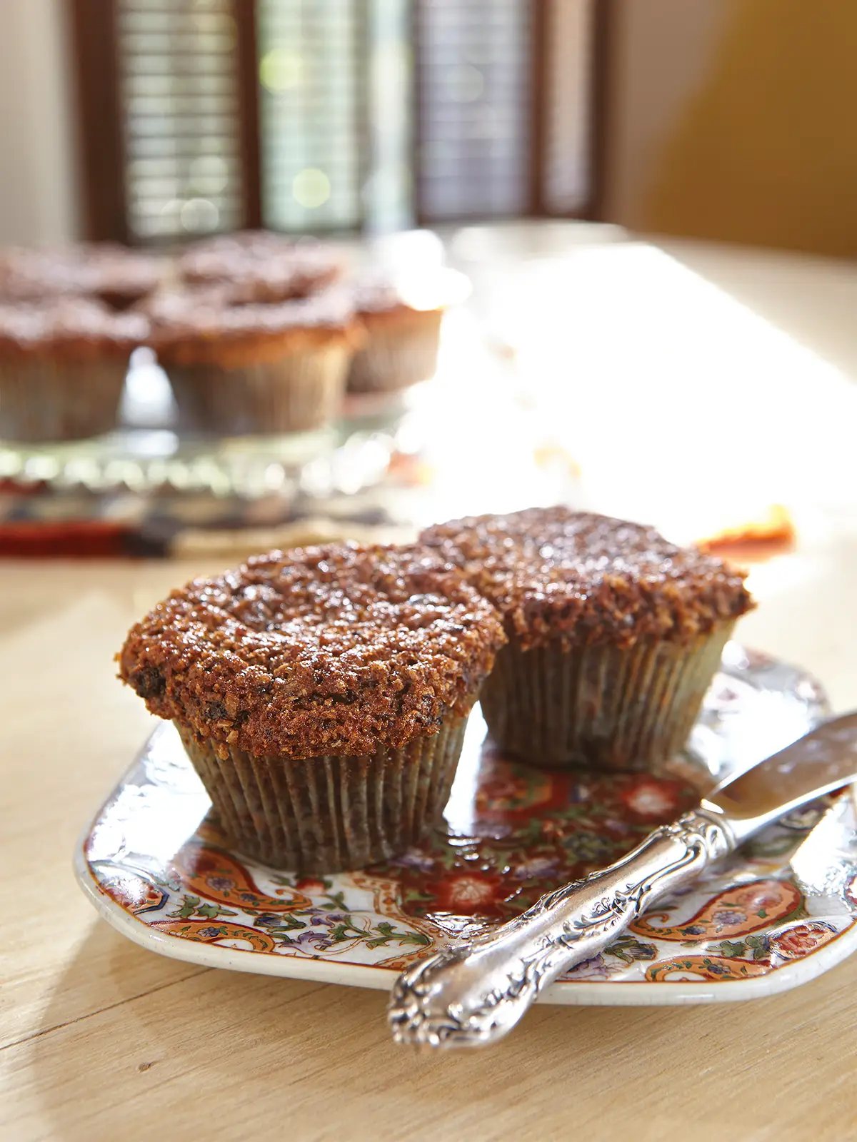 Two raisin bran muffins set on a patterned plate with a knife resting next to them.