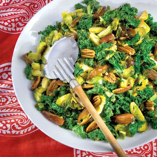 Roasted Kale, Brussels Sprouts, Date and Pecan Salad in a white bowl on a red patterned tablecloth.