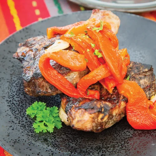 Roasted red peppers on top of grilled lamb chops on a round plate.