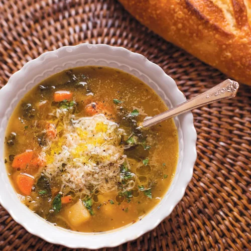 A bowl of winter minestrone and a baguette set on a round ratan placemat.