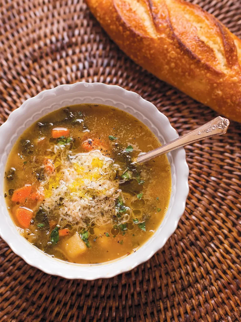 A bowl of winter minestrone and a baguette set on a round ratan placemat.