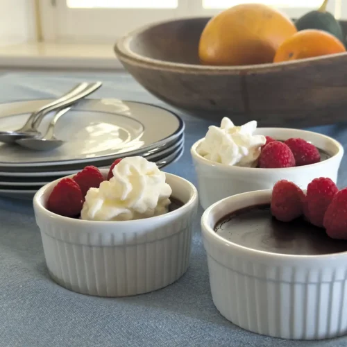 Three ramekins of Chocolate Coconut Pots de Crème on a blue tablecloth with a bowl of fruit behind them.