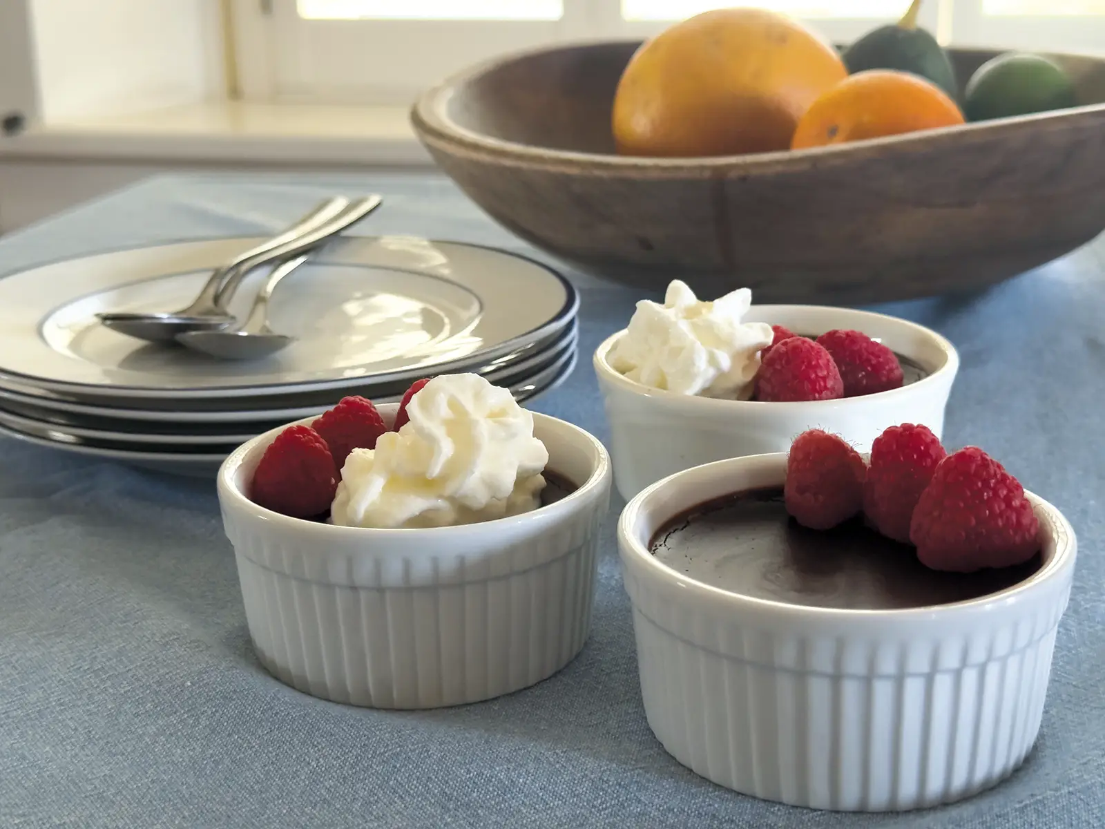 Three ramekins of Chocolate Coconut Pots de Crème on a blue tablecloth with a bowl of fruit behind them.