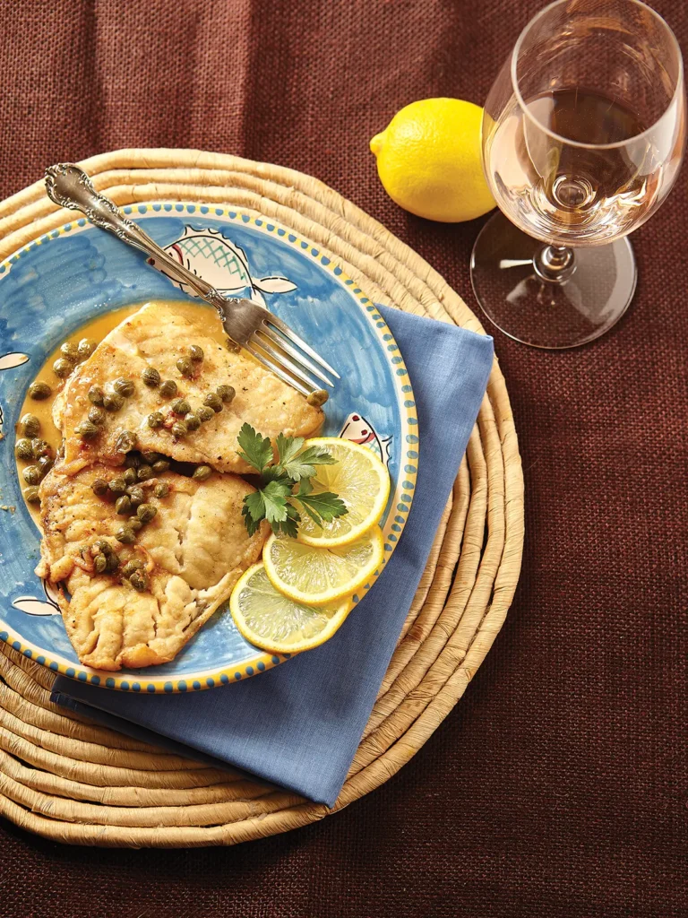 Tablescape with a plate of Lemony Fish Piccata on a round ratan placemat with a blue folded napkin, lemon, and a glass of wine.