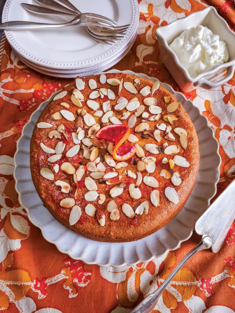 Orange almond cake on a white scalloped plate set on an orange tablecloth.