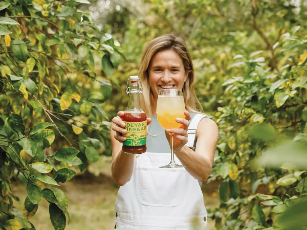 Sarah Bourke holding a bottle of Side Yard Vinegar in one hand and a glass of Side Yard Vinegar in the other.