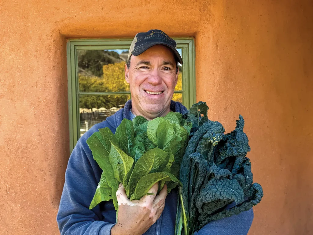 Sean Fennel standing against one of the buildings at Sunburst Sanctuary, holding bunches of greens in his arms.