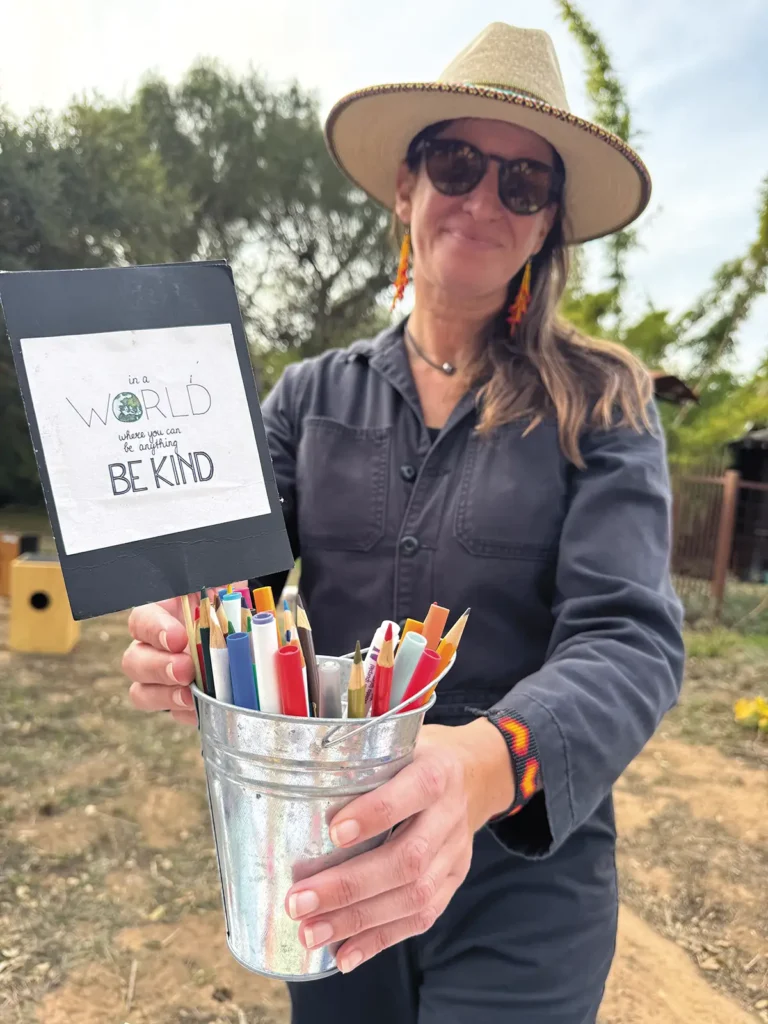 Sondra Weiss holds a bucket of colored pens and pencils with a sign that says "In a world where you can be anything, be kind"