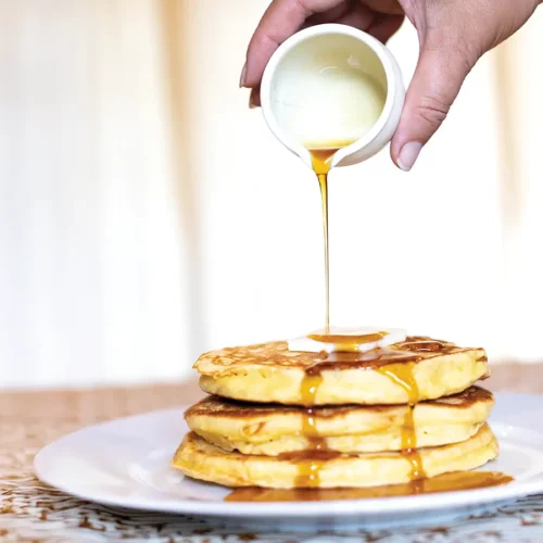Hand pouring syrup over a stack of pancakes with a pat of butter on top.