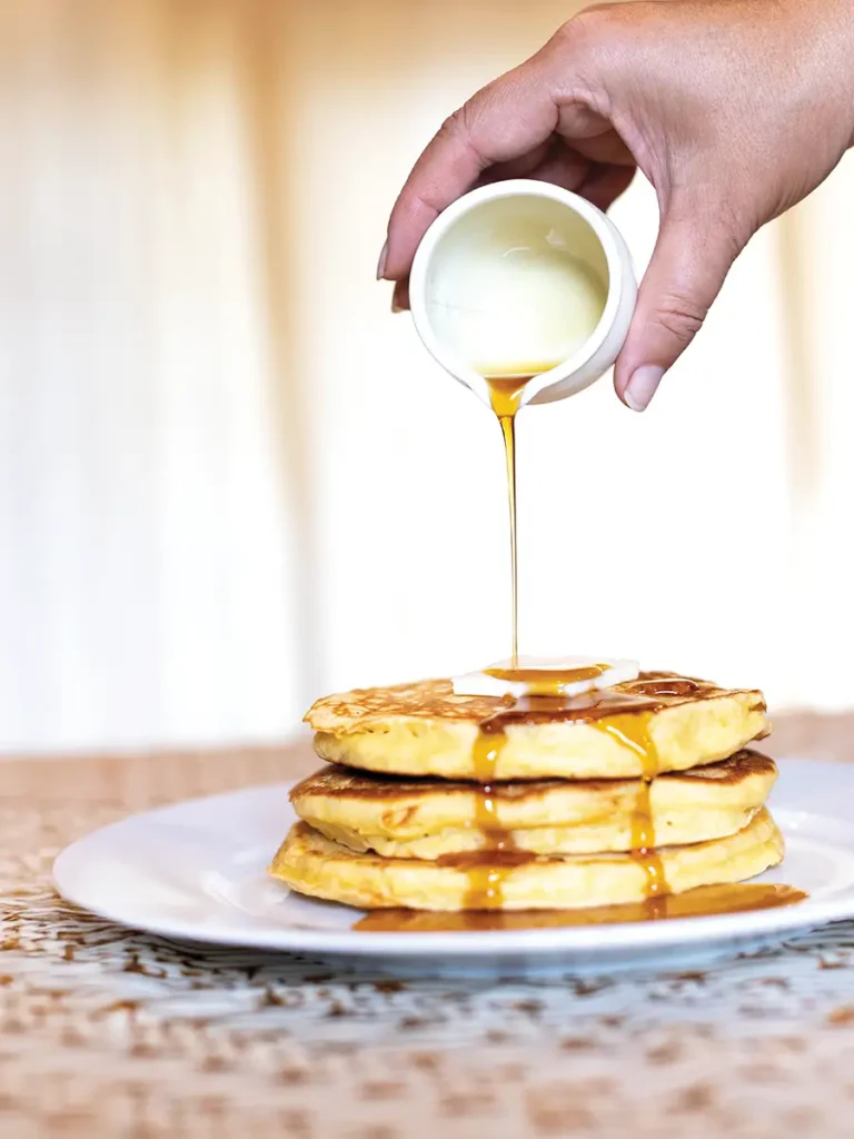 Hand pouring syrup over a stack of pancakes with a pat of butter on top.
