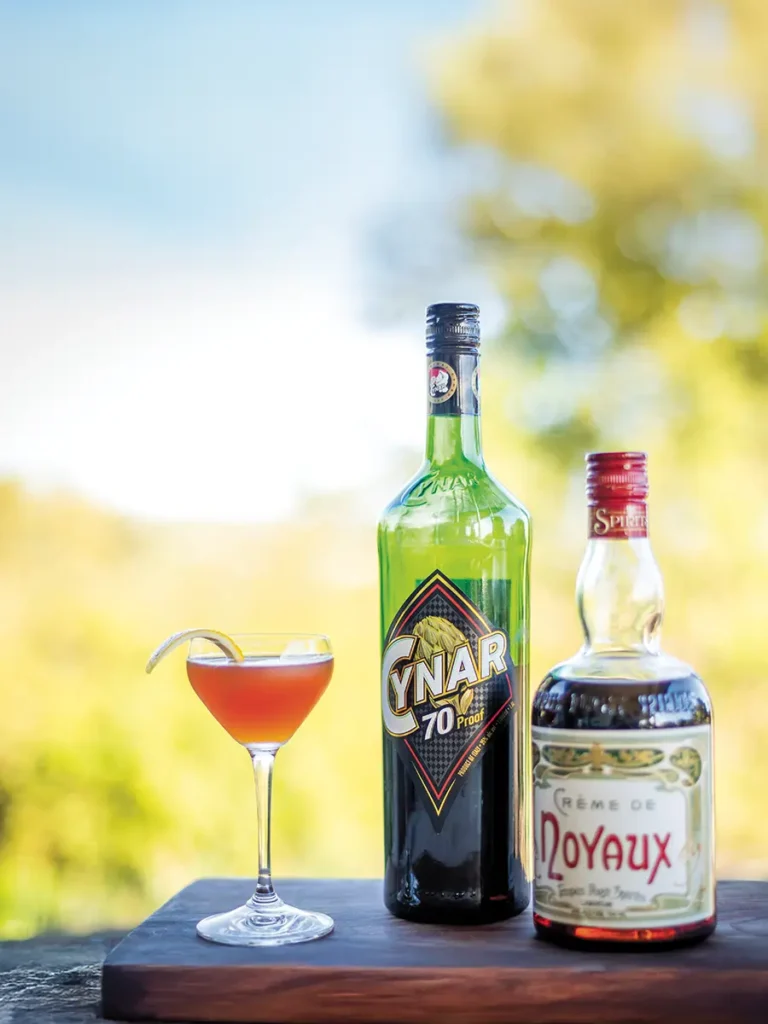 Pseudonymous Seed cocktail in a glass next to a bottle of Cynar and Crème de Noyaux.