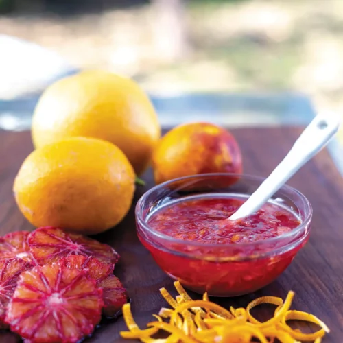 A bowl of marmalade sits on a wooden cutting board with sliced blood oranges and whole fruit.