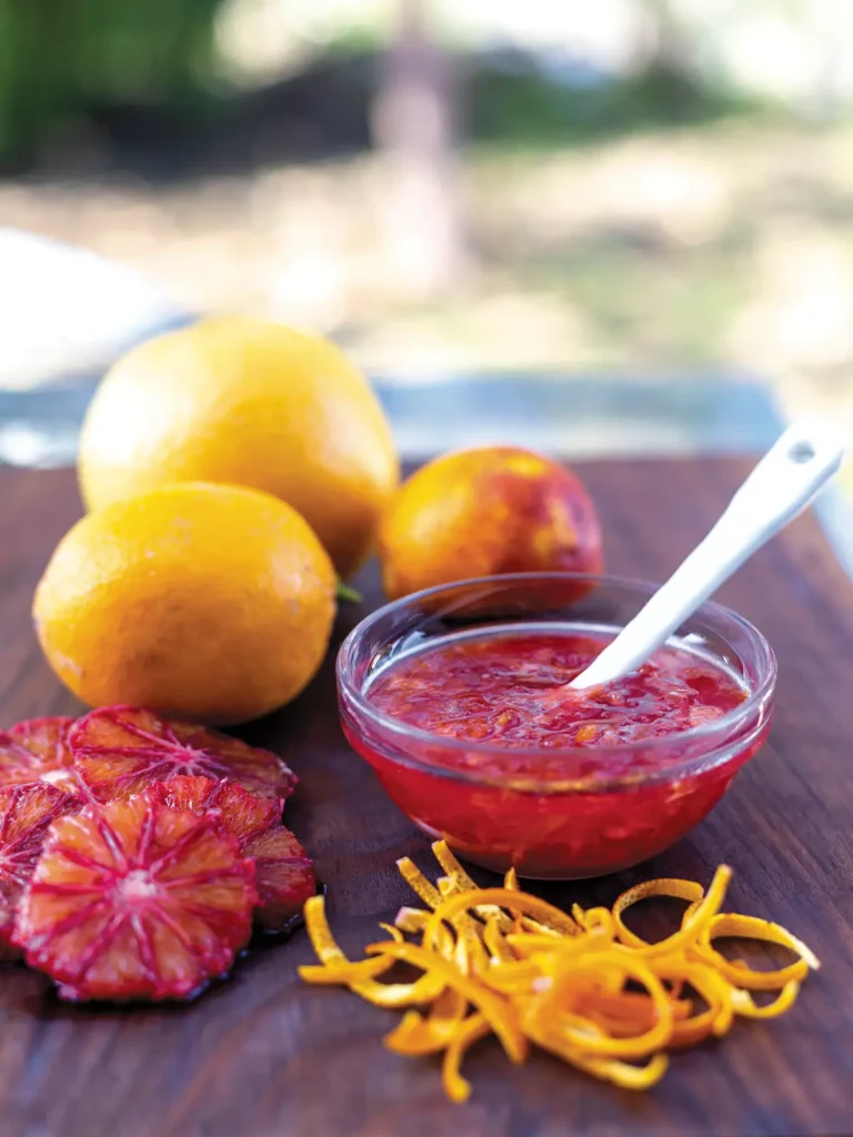A bowl of marmalade sits on a wooden cutting board with sliced blood oranges and whole fruit.
