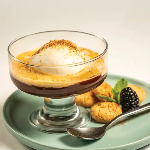 Clear coupe containing espresso and a scoop of gelato with Amaretti cookie crumbles on top. Cookies and a blackberry are set on a plate below the coupe along with a spoon.