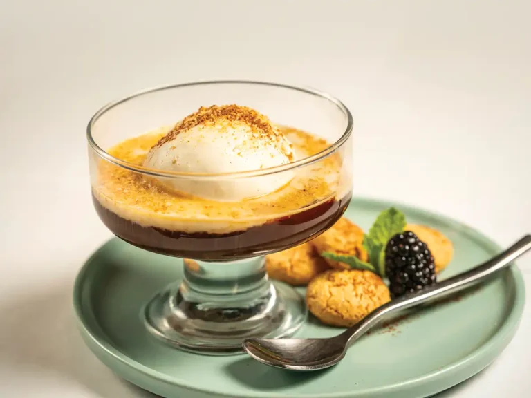 Clear coupe containing espresso and a scoop of gelato with Amaretti cookie crumbles on top. Cookies and a blackberry are set on a plate below the coupe along with a spoon.
