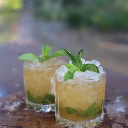 Two Myth Julep cocktails piled high with crushed ice and topped with mint sprigs on a wooden cutting board with ice scattered around it.