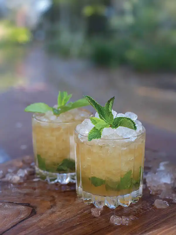 Two Myth Julep cocktails piled high with crushed ice and topped with mint sprigs on a wooden cutting board with ice scattered around it.