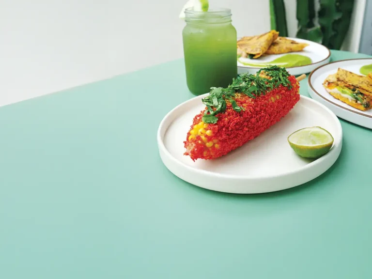 Spicy elote on a round white plate with a green agua fresca and some other dishes in the background.