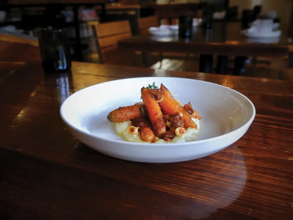 A white plate of roasted Roots Farm carrots with miso brown butter, parsnips, and hazelnuts.