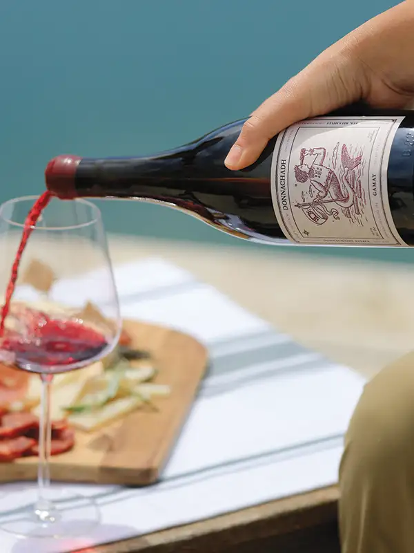 Someone pouring a glass of wine with a charcuterie board in the background.