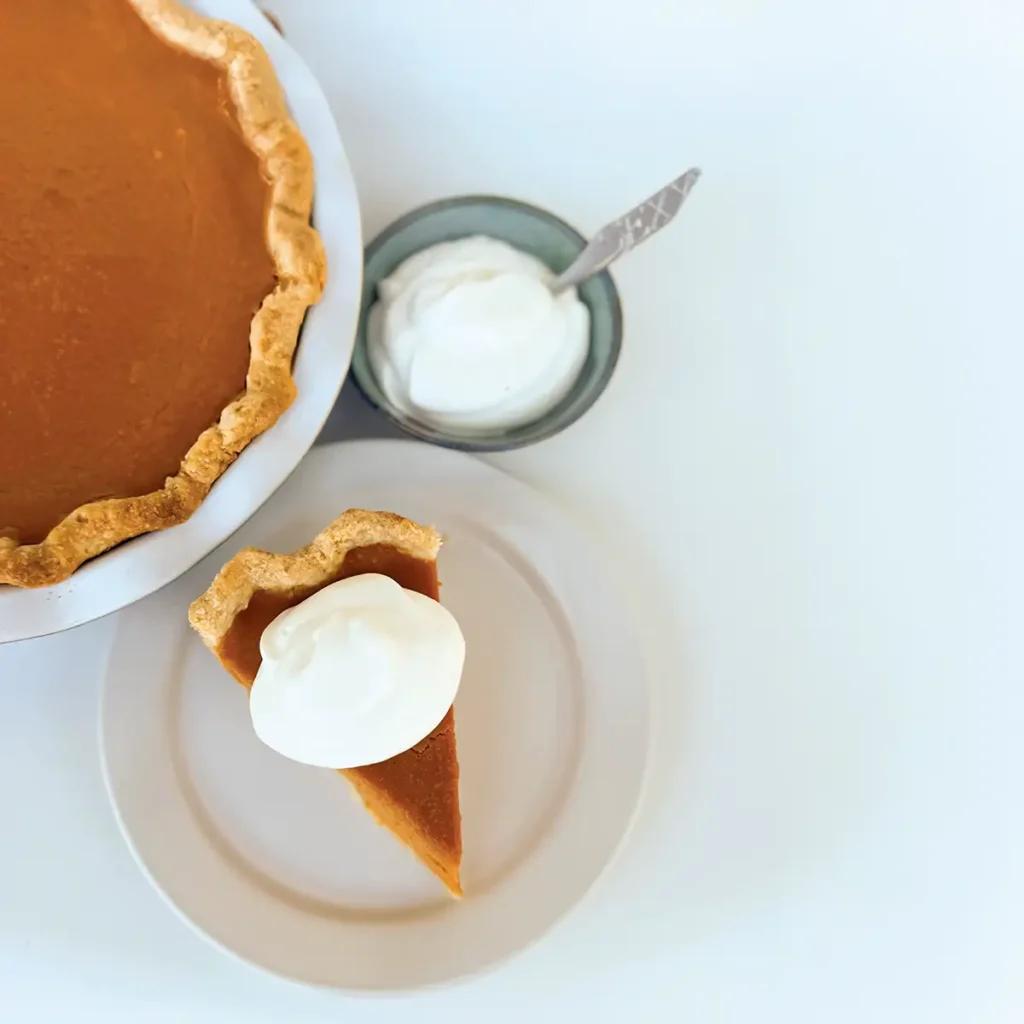 Pumpkin pie in a dish, a slice on a plate, and a bowl of whipped cream.