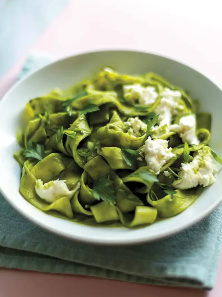 A white bowl with vibrant creamy kale pasta with mozzarella and herbs set on a dish towel.