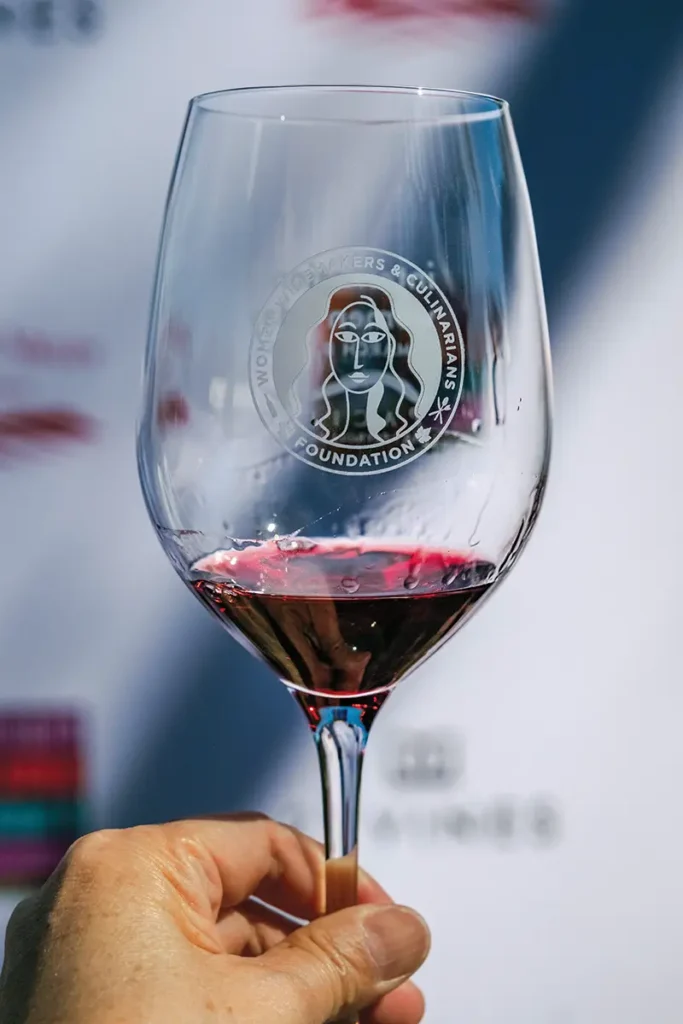 Hand holding a glass of red wine. The glass has the logo of the Women Winemakers & Culinarians Foundation.