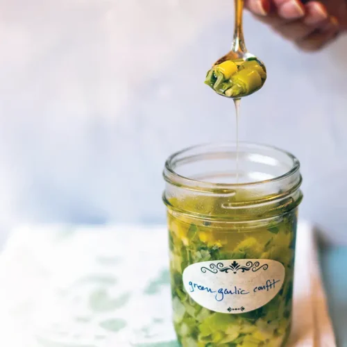 Hand holding a spoon of green garlic confit with the garlic oil streaming into a waiting mason jar below.