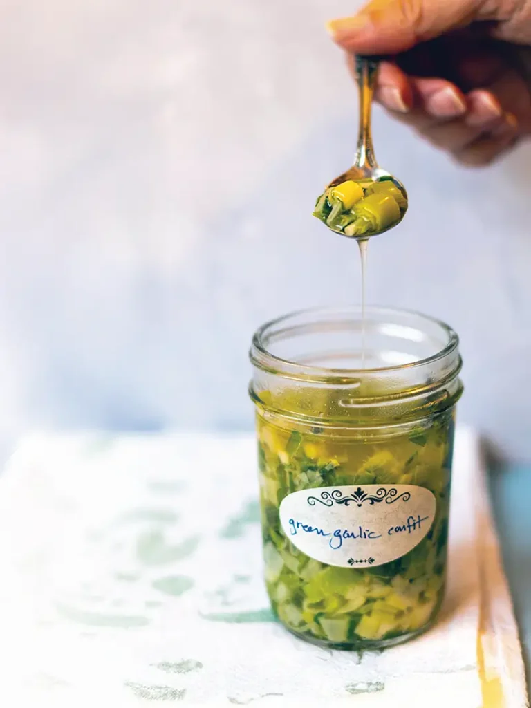 Hand holding a spoon of green garlic confit with the garlic oil streaming into a waiting mason jar below.