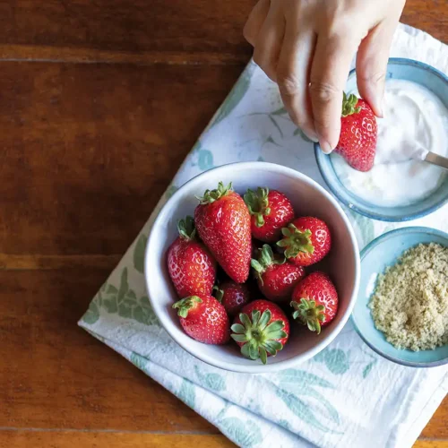 A bowl of fresh strawberries, a bowl of sour cream, and a bowl of brown sugar with a hand holding a strawberry ready to dip.