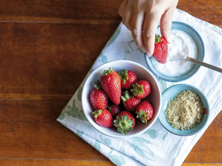 A bowl of fresh strawberries, a bowl of sour cream, and a bowl of brown sugar with a hand holding a strawberry ready to dip.