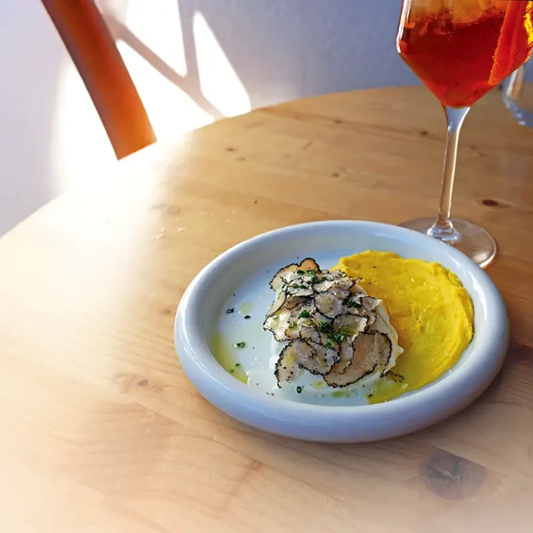 Aperitivo's Corn Pudding with Black Truffle and Buffalo Mozzarella is elegantly plated with delicate shaved black truffle, creamy buffalo mozzarella, and a vibrant corn purée, accompanied by a bright red Italian spritz.