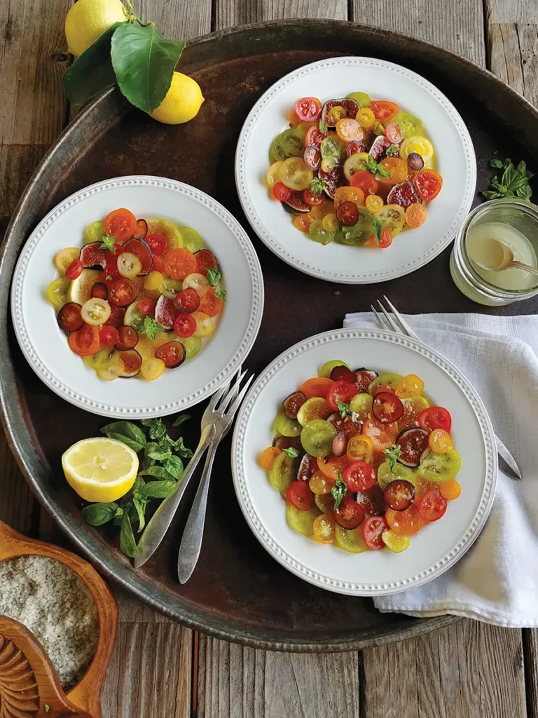 Three plates of colorful sliced Fig and Cherry Tomato Mosaic with Goat Cheese Herb Crema arranged on a rustic metal tray.