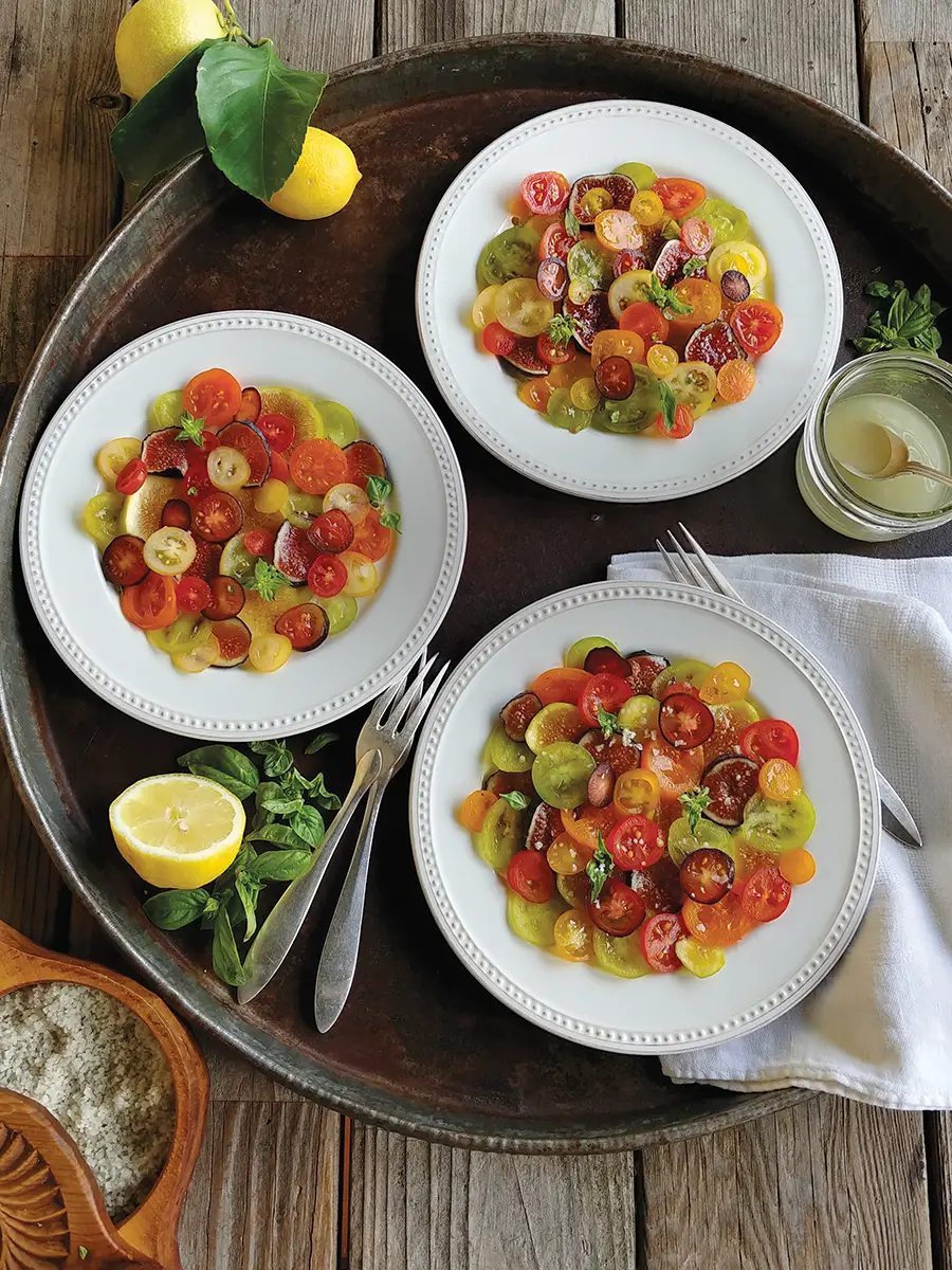 Three plates of colorful sliced Fig and Cherry Tomato Mosaic with Goat Cheese Herb Crema arranged on a rustic metal tray.