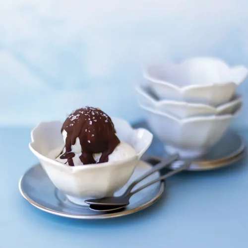 Vanilla ice cream in a white scalloped bowl topped with glossy chocolate sauce and coarse salt, served with spoons on a pale blue table.