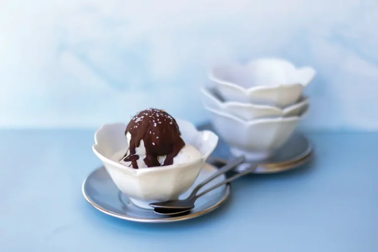 Vanilla ice cream in a white scalloped bowl topped with glossy chocolate sauce and coarse salt, served with spoons on a pale blue table.