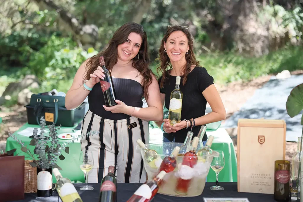 Two women smiling at an outdoor wine tasting table, holding bottles of wine with chilled rosé and white wines displayed in an ice bucket.