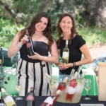 Two women smiling at an outdoor wine tasting table, holding bottles of wine with chilled rosé and white wines displayed in an ice bucket.