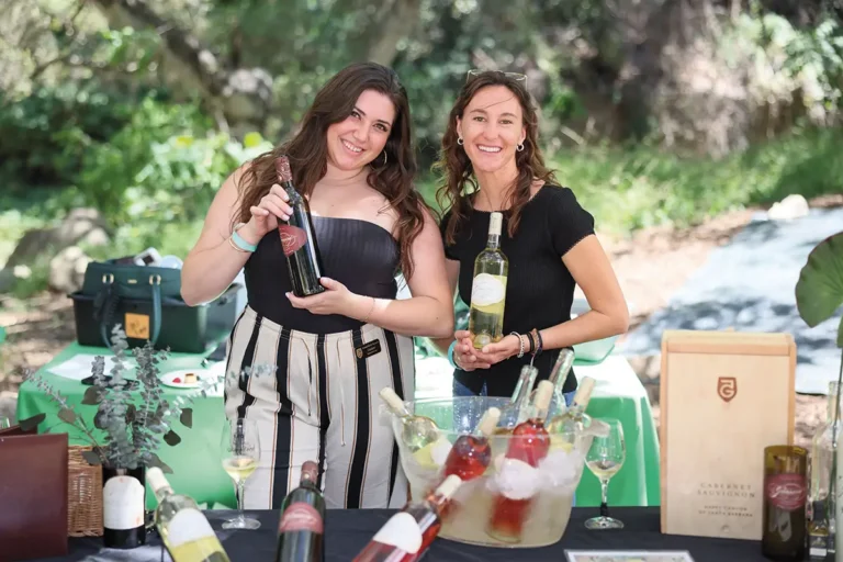 Two women smiling at an outdoor wine tasting table, holding bottles of wine with chilled rosé and white wines displayed in an ice bucket.